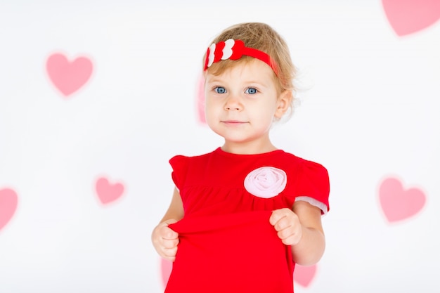 Little blonde girl in red dress with red wreath with hearts on white with pink hearts on the  Valentines day