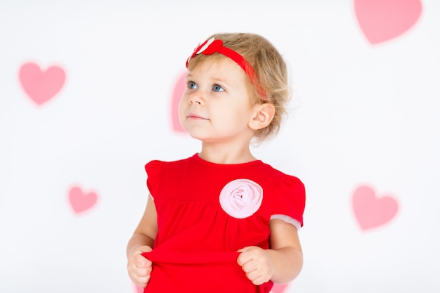 Little blonde girl in red dress with red wreath with hearts on white with pink hearts on the  Valentines day