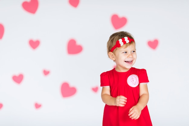 Little blonde girl in red dress with red wreath with hearts on white with pink hearts on the  Valentines day