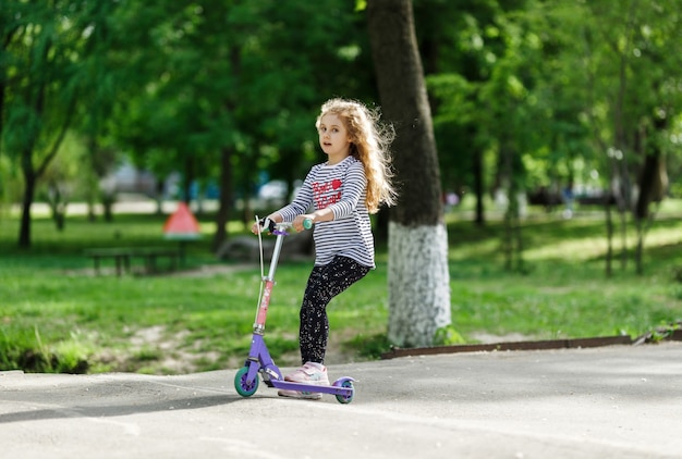 小さなブロンドの女の子が公園でスクーターに乗る。