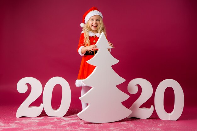 サンタのスーツの金髪少女は大きな白いクリスマスツリーを保持しています。