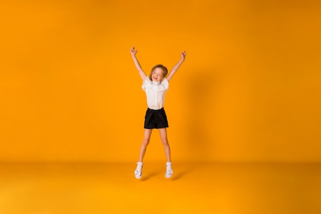 a little blonde girl in a school uniform jumps on a yellow background 