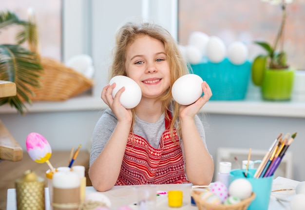 Little blonde girl showing eggs before coloring for Easter holiday at home.