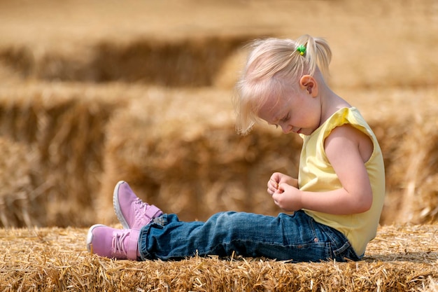 小さなブロンドの女の子が干し草の中に座っています。子供は干し草の山を再生します。農場での子供時代。夏の日。