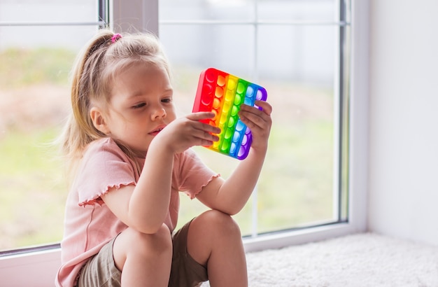 小さなブロンドの女の子が窓の近くに座って、新しいトレンドの感覚玩具で遊んでいます-レインボーポップ。 AntistressÃƒÂ'Ã‚Â olorfultoyシンプルなディンプル。スクイーズソフトバブルおもちゃレインボーカラー