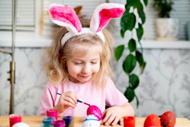 A little blonde girl sits at the table and paints easter eggs with brushes and paints smiles