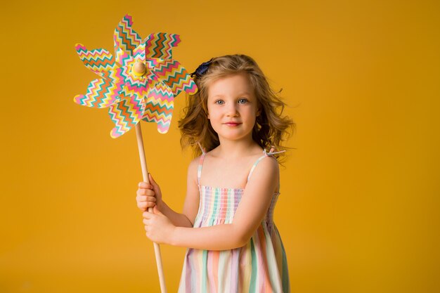 Little blonde girl smiles in a summer dress, sunglasses, holding a child's windmill on a yellow wall