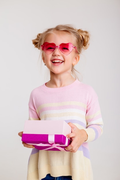 Bambina bionda che sorride in occhiali da sole a forma di cuore rosa che tengono un contenitore di regalo rosa su un fondo bianco