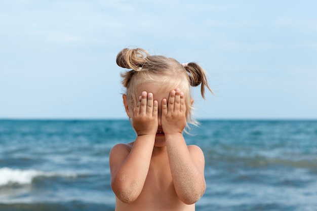 小さなブロンドの女の子は海の背景に立って、彼女の手で彼女の顔を覆います