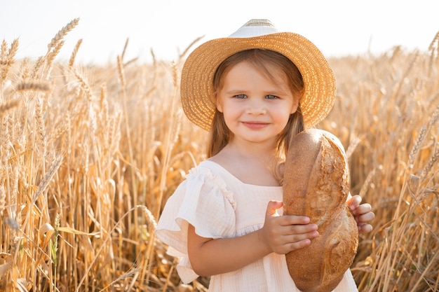 Photo little blonde girl in straw hat and beige muslin dress holds bread in wheat field on sunset