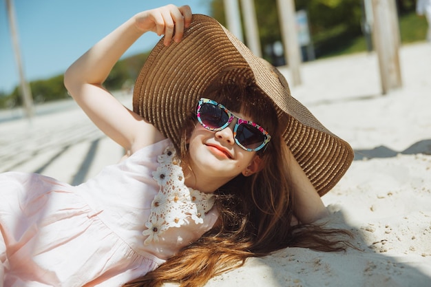 Photo a little blonde girl in a straw hat lies on a sandy beach girl sunbathing on the sand