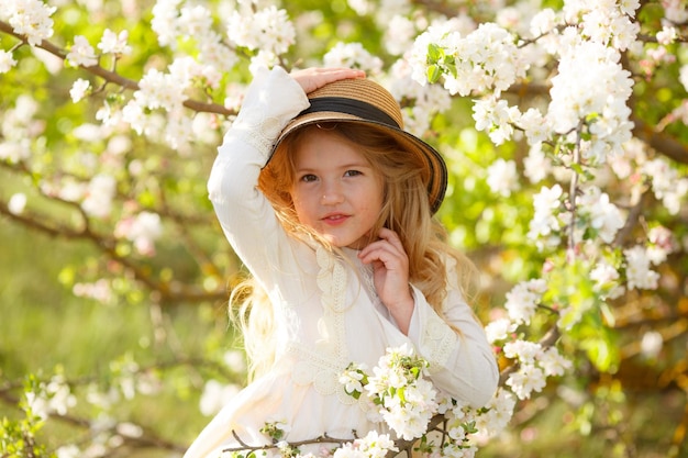 A little blonde girl in a straw hat near a flowering tree