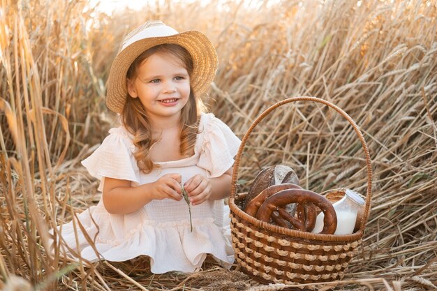 Little blonde girl in straw hat with basket of bread in wheat field. eco friendly farm products.