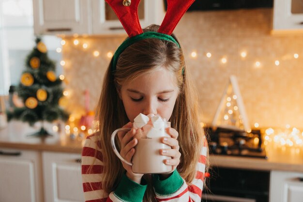 La piccola ragazza bionda in pigiama a righe tiene una tazza con una bevanda calda e marshmallow nella cucina decorata. fine settimana e festivi invernali. posto per il testo.