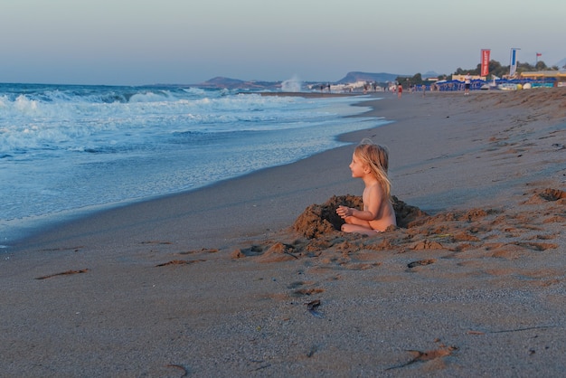 Una piccola ragazza bionda in costume da bagno si siede su una spiaggia sabbiosa in riva al mare e gioca con la sabbia a