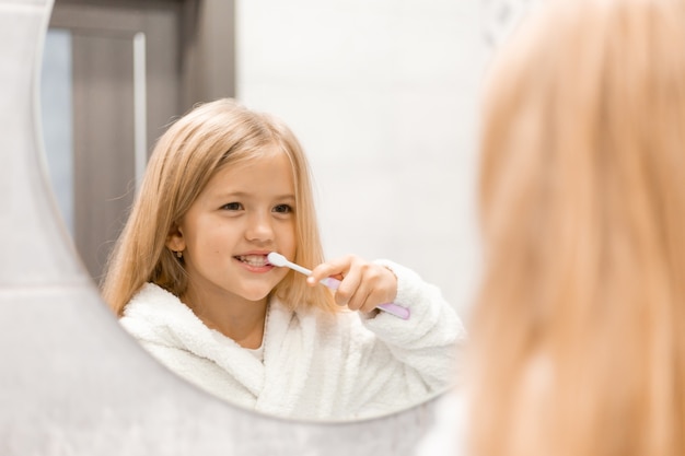 little blonde girl in a white bathrobe brushes her teeth in front of the bathroom mirror