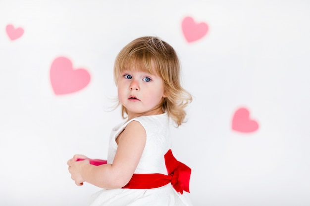 Piccola ragazza bionda in abito bianco con nastro rosso sul pavimento bianco con cuori rosa il giorno di san valentino