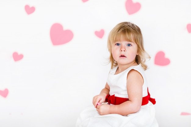 Piccola ragazza bionda in abito bianco con nastro rosso sul pavimento bianco con cuori rosa il giorno di san valentino