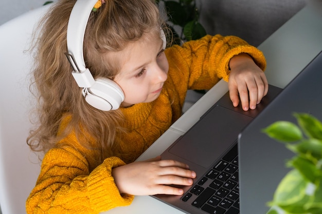 A little blonde girl in white headphones and a yellow sweater learns at a laptop remote learning online lesson