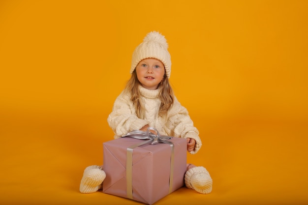 a little blonde girl in a white knitted sweater hat and socks with a gift box in her hands