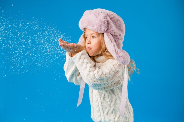 冬の服を着た小さなブロンドの女の子は彼女の手から雪を吹きます