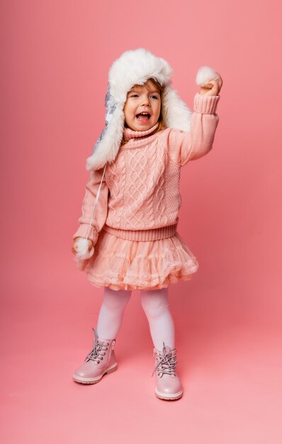 冬の服とピンクの背景に毛皮の帽子の小さなブロンドの女の子。