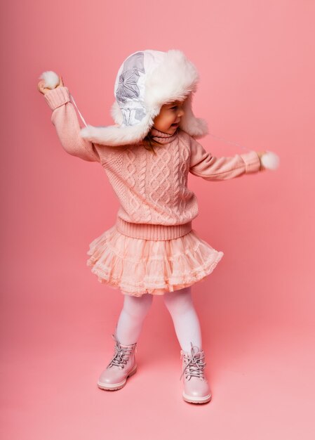 Bambina bionda in abiti invernali e un cappello di pelliccia su uno sfondo rosa.