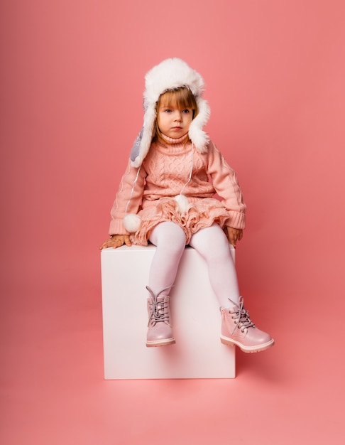 Little blonde girl in winter clothes and a fur hat on a pink background.