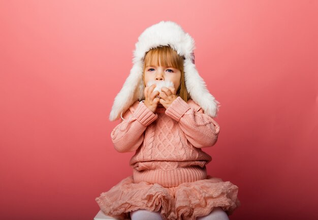 Little blonde girl in winter clothes and a fur hat on a pink background.