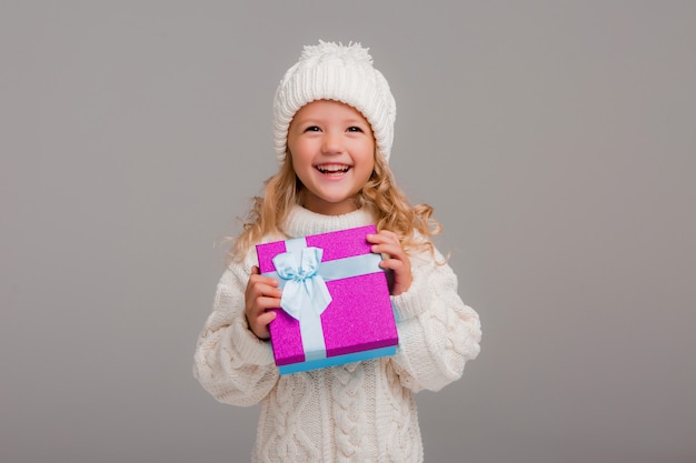 Piccola ragazza bionda in cappello di inverno che sorride tenendo un contenitore di regalo