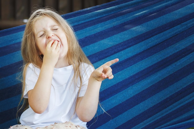 長い髪の小さなブロンドの女の子は、青いハンモックに座って庭でイチゴを食べる