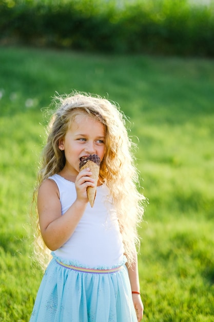 Little blonde girl with long hair has fun eating ice cream in a summer park. 