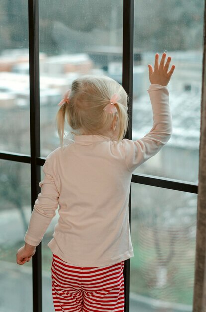 Little blonde girl with two ponytails in red striped pants stands near large window and waits for someone Vertical frame