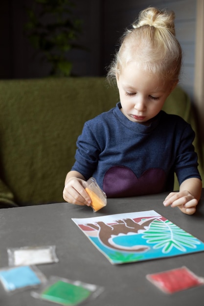 Little blonde kid girl creating sand picture of dinosaur leisure activity indoors creative hobby