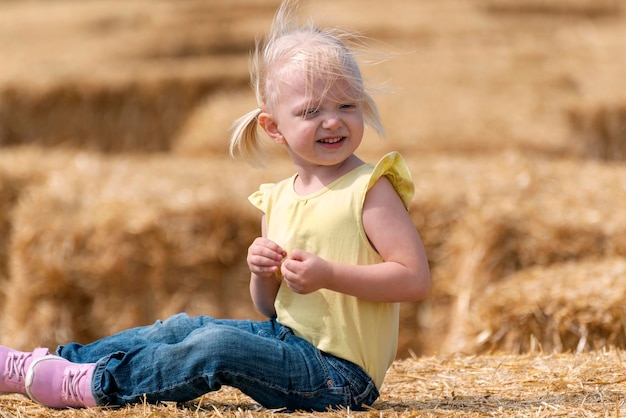 Little blonde smiling girl sits in the hay. Happy child plays haystack. Childhood on the farm. Summer day.