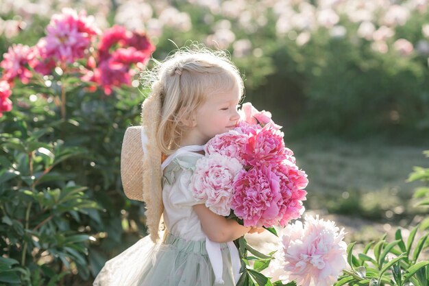 Foto bambina contadina bionda in abito e cappello di paglia in peonie in fiore primavera