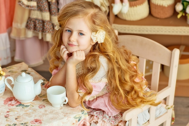 A little blonde with long hair smiles a beautiful girl 6 years\
old in a retro sitting at a table and drinking tea.baby chic\
style