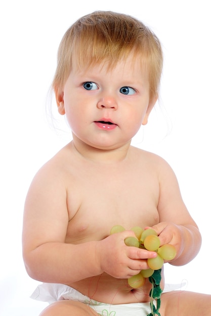 Piccolo neonato degli occhi azzurri che si siede vicino al canestro di vimini e che tiene l'uva nelle mani