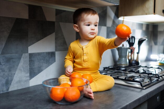 黄色の服を着た 1 歳の男の子がテーブルの上のキッチンに座って、オレンジ色のみかんのプレートを持っています かわいい 1 歳の男の子と甘い柑橘系の果物の肖像画