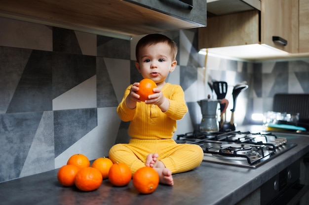 黄色の服を着た 1 歳の男の子がテーブルの上のキッチンに座って、オレンジ色のみかんのプレートを持っています かわいい 1 歳の男の子と甘い柑橘系の果物の肖像画