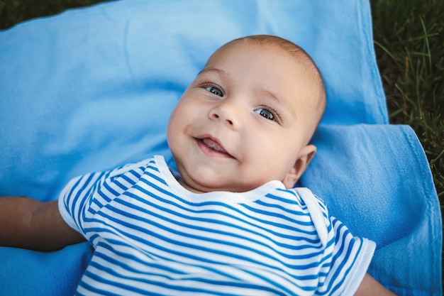 A little boy of 3 months lies on his back on a blue bedspread in the park around the green grass and trees. Children's emotions of joy