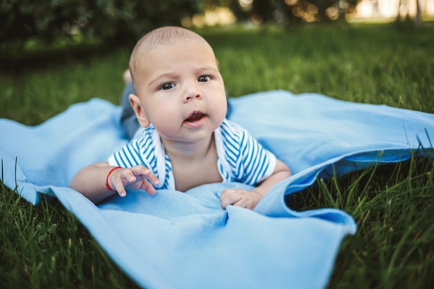 Little boy 3 months old lies on his stomach on a blue bedspread in the park around the green grass and trees. Children's emotions of joy