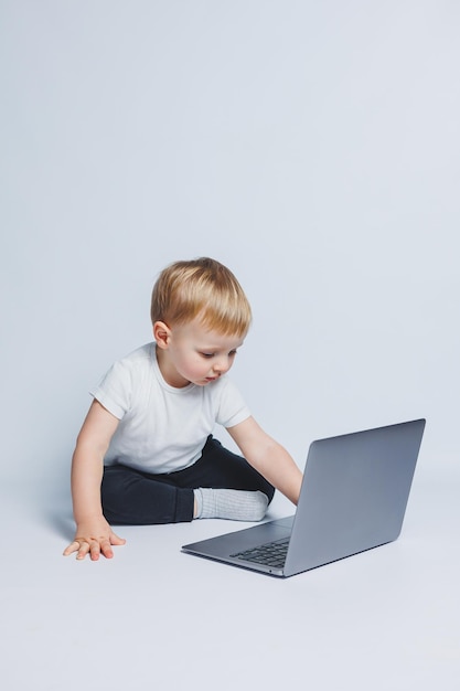 A little boy 34 years old sits with a laptop on a white background A child in a white Tshirt and black trousers looks at a laptop Modern children