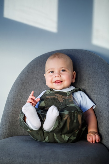 Маленький мальчик 8 месяцев в комбинезоне, белой рубашке и белых носках сидит на сером стуле