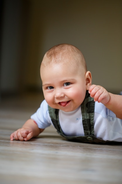 Little boy aged 8 months in overalls, a white shirt and white socks crawling on the floor and smiling