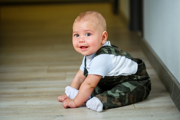 Little boy aged 8 months in overalls, a white shirt and white socks crawling on the floor and smiling