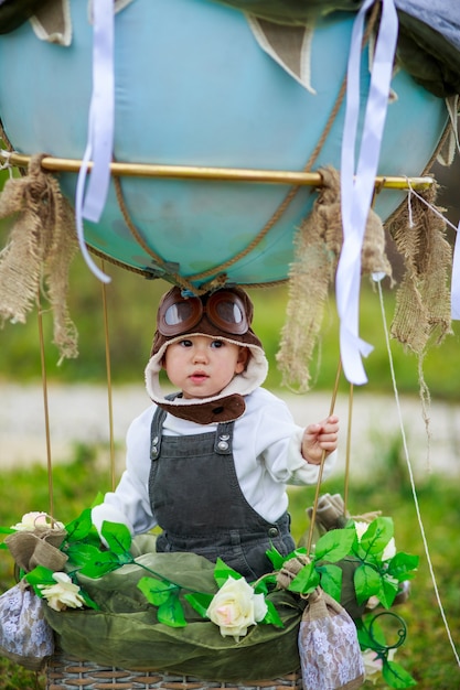 A little boy in a aviator's hat sits in a basket of a toy balloon in a field.