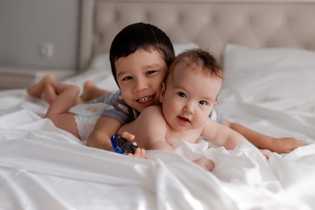 little boy and a baby two brothers are lying on white bed linen in bed and hugging