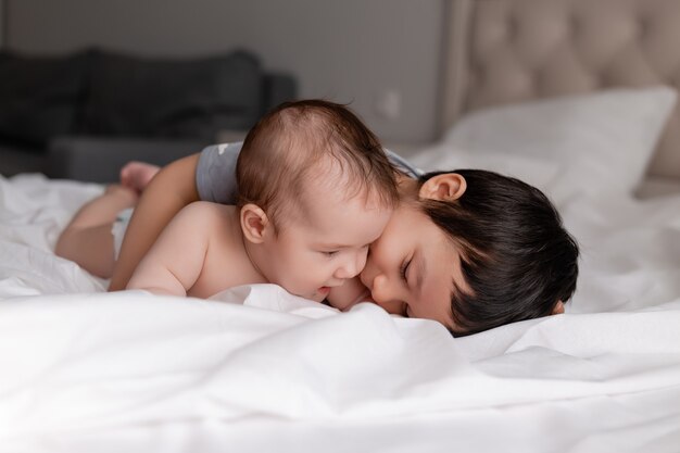 little boy and a baby two brothers are lying on white bed linen in bed and hugging