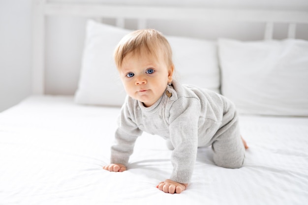 Маленький мальчик блондинка с голубыми глазами в сером костюме на четвереньках дома в спальне на кровати с белым бельем смотрит в камеру